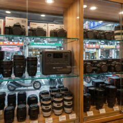 二手相機店的玻璃展示櫃中，除了單眼相機與鏡頭外，還陳列著一台 DJI Power 1000，象徵多元收購 (Inside a used camera store display case, alongside DSLR cameras and lenses, a DJI Power 1000 is displayed, symbolizing diverse buyback options).