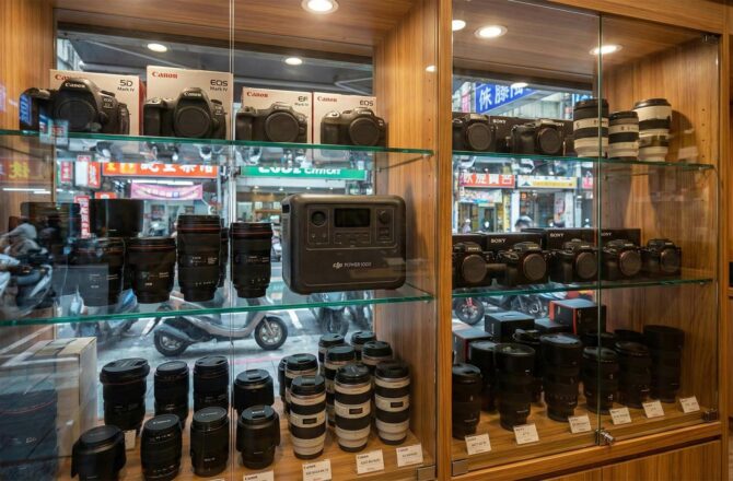 二手相機店的玻璃展示櫃中，除了單眼相機與鏡頭外，還陳列著一台 DJI Power 1000，象徵多元收購 (Inside a used camera store display case, alongside DSLR cameras and lenses, a DJI Power 1000 is displayed, symbolizing diverse buyback options).