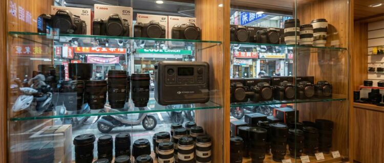 二手相機店的玻璃展示櫃中，除了單眼相機與鏡頭外，還陳列著一台 DJI Power 1000，象徵多元收購 (Inside a used camera store display case, alongside DSLR cameras and lenses, a DJI Power 1000 is displayed, symbolizing diverse buyback options).